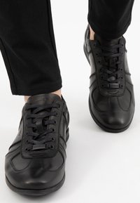 Černé kožené tenisky s kulatou špičkou, šněrovacím designem, texturovaným svrškem a decentním logem. V kombinaci s černými kalhotami.