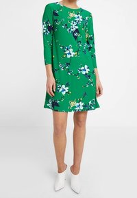 Green dress with three-quarter sleeves, featuring a floral pattern in white, blue, and yellow. A-line shape, knee-length. Worn with white heels.