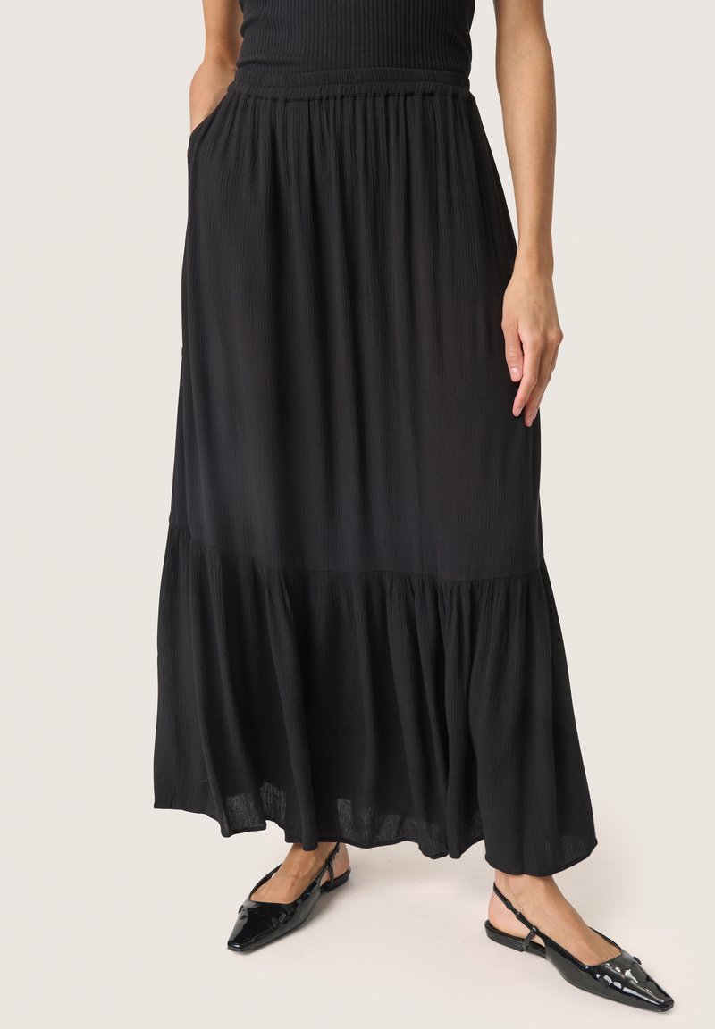 Jupe maxi noire à volants en tissu texturé, avec une taille élastique et des poches latérales, associée à des ballerines noires à bout pointu.