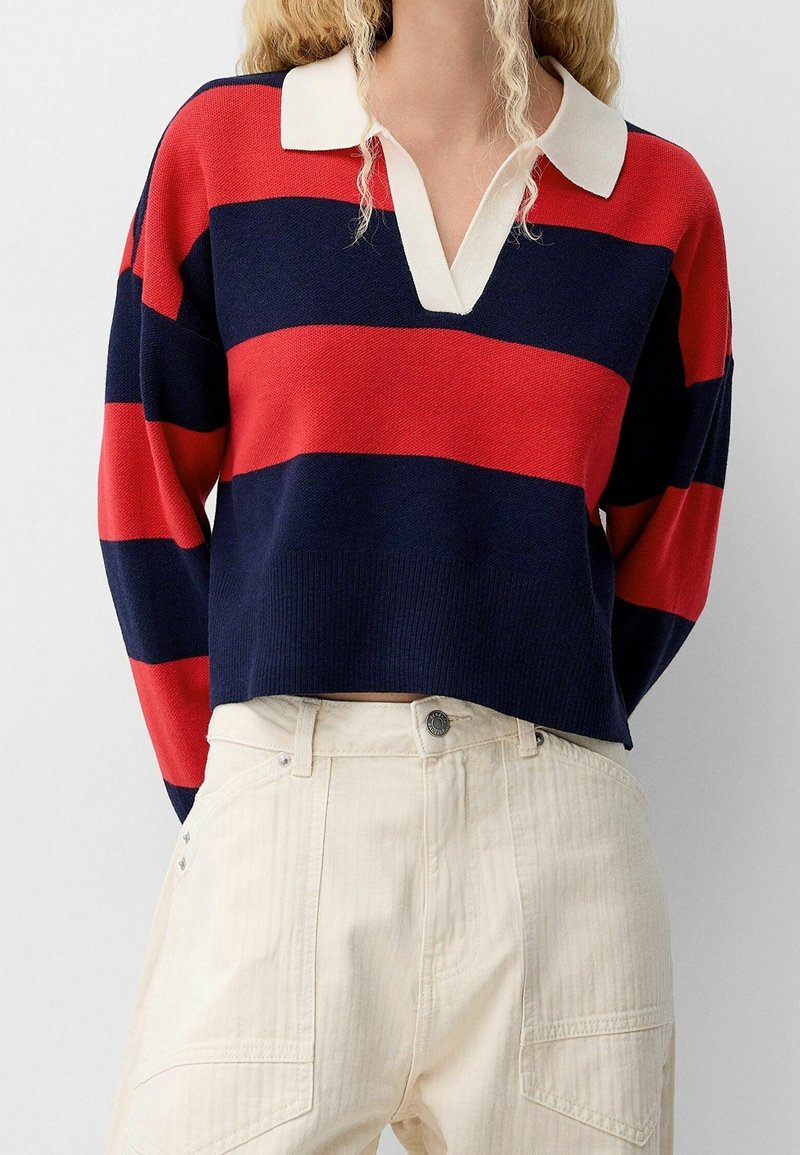 Pull rayé rouge et marine avec un col blanc, présentant une coupe décontractée et une texture côtelée. Associé à un pantalon crème taille haute.