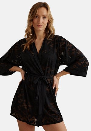 Jeune femme portant une robe portefeuille noire transparente à larges manches, nouée à la taille, posant les mains sur les hanches devant un fond blanc.