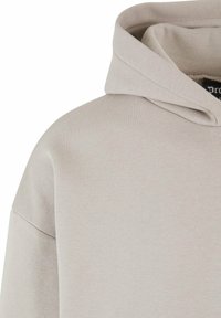 Haut à capuche beige clair au toucher doux, doté d'une grande capuche et d'une coupe décontractée. Coutures plates et détails de conception minimaux sont présents.