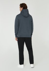 Hoodie i mörkgrått tyg med en avslappnad passform, visad från baksidan, ihopsatt med svarta byxor och vita sneakers.