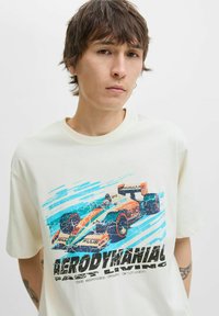 Tricou de culoare crem, cu un grafic al unei mașini de curse în nuanțe de portocaliu și albastru, și cu un text negru îndrăzneț care citește "AERODINAMICĂ" sub imagine.