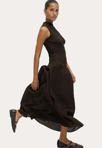 Vrouw in een mouwloze zwart gekleurde jurk met een volle, vloeiende rok, die zwarte platte Mary Jane schoenen draagt en in profiel poseert.