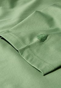 Világos zöld, puha tapintású pamut anyag, amelyen a hajtott ujján egy kicsit sötétebb zöld színű hímzett logó található.