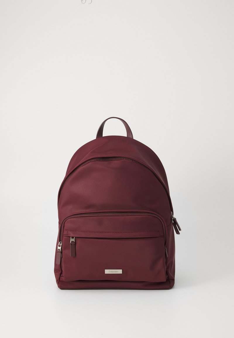 Κερασί backpack από νάιλον με στρογγυλό πάνω μέρος, μπροστινή τσέπη με φερμουάρ και χερούλι στην κορυφή. Διαθέτει ασημί μεταλλικά στοιχεία και πλακέτα λογοτύπου στο μπροστινό μέρος.