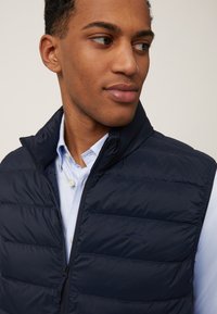 Mornarska pufasta prsluk s visokim ovratnikom, zatvaranjem na munju i horizontalnim šavovima. Nositi preko svijetloplavog košulje s ovratnikom. Suptilan detalj loga.