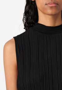 Haut sans manches noir avec un col montant, présentant des plis verticaux et une texture lisse. Design simple sans motifs ni embellissements visibles.