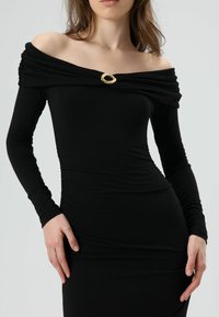 Schwarzes Langarmoberteil mit schulterfreiem Design, ausgestattet mit drapiertem Stoff und einem goldenen ovalen Broschendetail am Ausschnitt.