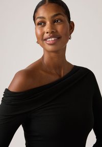 Μαύρο τοπ με ανοιχτούς ώμους από απαλό ύφασμα, με πτυχώσεις και εφαρμοστή γραμμή. Το μοντέλο φορά μικρούς ασημένιους κρίκους σκουλαρίκια.