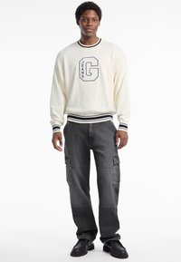 Suéter crema con puños a rayas azul marino, que presenta un gran bordado de "G" y "JEANS". Pantalones cargo negros con bolsillos laterales y un corte holgado.