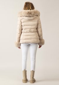 Beige Daunenjacke mit fellbesetztem Kragen, tailliertem Schnitt und gepufften Ärmeln. Getragen über weißen Skinny-Jeans und beigen Stiefeln. Weicher Stoff, gestepptes Design.