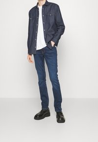 Camisa de mezclilla oscura con botones a presión, camiseta blanca debajo, vaqueros ajustados azul oscuro y zapatos de plataforma negros. Textura de tela suave.