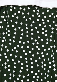 Tela verde con lunares blancos de diferentes tamaños. Presenta una textura suave y un diseño simple con una sección fruncida en la parte superior.