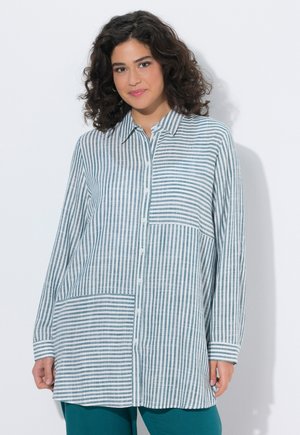 Kiharaiset tummatukkaiset nainen pukeutuneena väljään valkoiseen paitaan, jossa on vaihtelevia sinisiä pysty- ja vaakaraitoja, seisoo valkoista taustaa vasten.