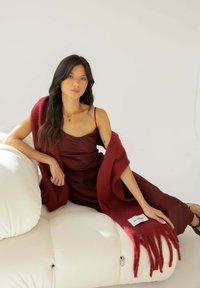 Burgunderfarbige Wolldecke mit fransigen Rändern, die über ein elegantes, dunkles Kleid gelegt ist, das auf einem weißen, kuscheligen Sofa mit sichtbaren Nähten ruht.