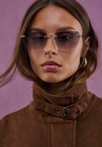 Veste en suède marron avec un col montant et des détails de boucle, associée à des lunettes de soleil surdimensionnées grises avec une monture transparente.