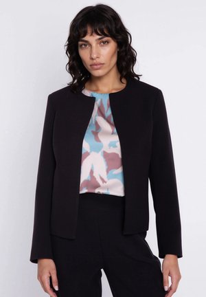 Kobieta o ciemnych włosach do ramion, ubrana w czarną, otwartą kurtkę na abstrakcyjnie wzorzystej bluzce w kolorach niebieskim, różowym i białym, pozująca na jasnym tle.