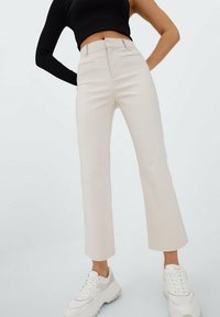 Crème kleurige, high-waisted, flare broeken met voorzakken en een gladde textuur, gecombineerd met witte sneakers.