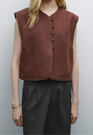 Femme portant un gilet marron sans manches boutonné et un pantalon plissé gris foncé taille haute avec un bracelet doré au poignet gauche.