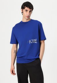 Karališkai mėlynas medvilnės marškinėlis, pasižymintis apvalia iškirpte ir trumpomis rankovėmis, priekyje su baltu ir auksiniu logotipu.