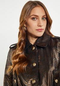 Zwarte en metallic slangenpatronen jas met een kraag, gouden knopen en een glanzende afwerking. Lange, golvende bruine haren zichtbaar.