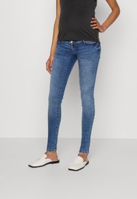Schmale Jeans aus mittlerem blauen Denim mit einer mittelhohen Taille, fünf Taschen und einem dunkel gefärbten Oberteil. Weiße Slipper runden das Outfit ab.