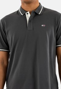 Polo de color gris oscuro con una superficie texturizada. Presenta detalles en blanco en el cuello y las mangas. Pequeño logo en la zona del pecho izquierdo.