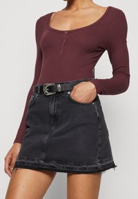 Kvinna i en figurnära vinröd långärmad topp med knappar, samt en svart denim minikjol med svart bälte och silverspänne.