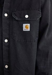 Black denim shirt with silver buttons, two chest pockets, and a white square patch featuring a yellow logo. Textured fabric, casual design.