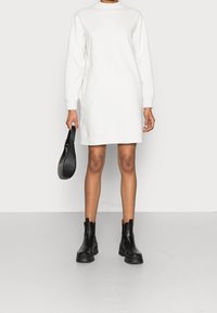 Abito felpa bianco con maniche lunghe, collo alto e vestibilità dritta. Abbinato a stivali neri alla caviglia e una piccola borsa nera.