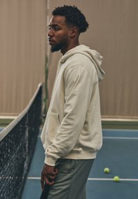 Beige hoodie with a front pocket and drawstring hood, paired with light gray sweatpants. Tennis court net and balls visible in the background.