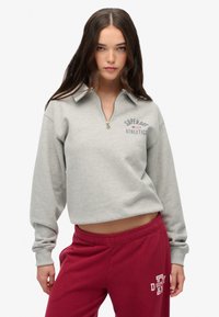 Sudadera gris corta con cuello de media cremallera y logo bordado "Superdry Athletics". Lleva puestas pantalones de chándal burdeos con texto blanco.