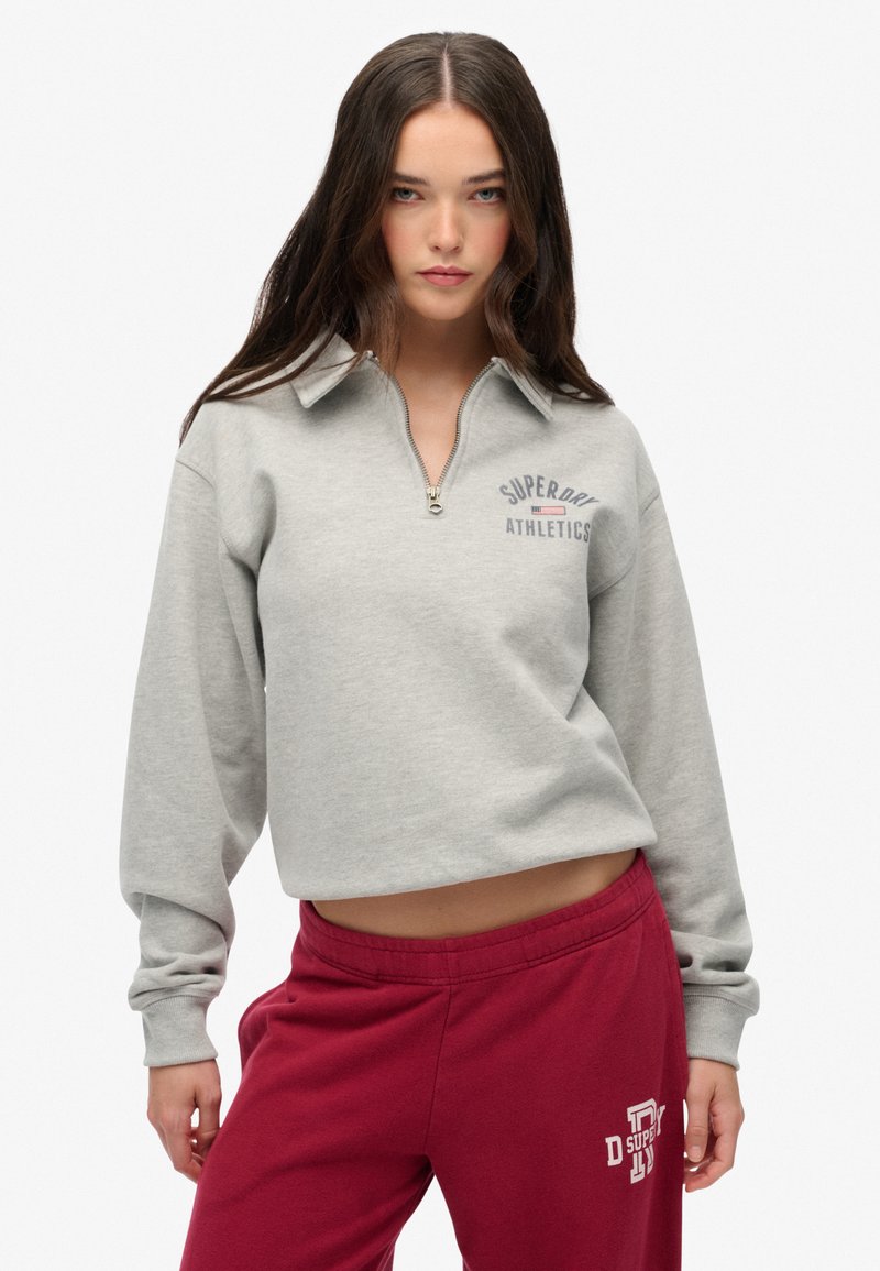 Sudadera gris corta con cuello de media cremallera y logo bordado "Superdry Athletics". Lleva puestas pantalones de chándal burdeos con texto blanco.