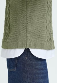 Groene gebreide trui met een textuurpatroon over een wit overhemd met kraag, gecombineerd met donkere spijkerbroek. Zichtbare ribbels aan de onderkant en gelaagd ontwerp.