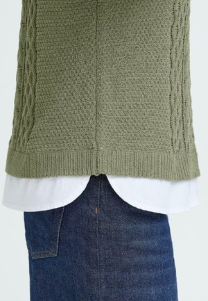 Jersey verde de punto con un patrón texturizado sobre una camisa blanca de cuello, combinado con jeans de mezclilla oscura. Puños ribeteados visibles y diseño en capas.