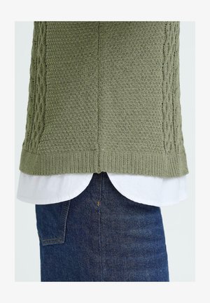 Grönstickad tröja med ett texturerat mönster över en vit skjorta med krage, kombinerad med mörka denimjeans. Synliga ribbade nederkanter och lagerdesign.