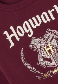 Sweatshirt bordeaux avec un imprimé blanc "Hogwarts", un logo de blason avec des accents dorés et des graphiques de couronnes de laurier décoratives.