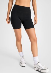 Pantalones cortos de ciclismo negros de talle alto, fabricados en un tejido elástico, con una textura suave y un diseño ajustado, combinados con zapatillas blancas y calcetines.