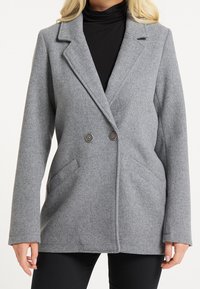 Manteau en mélange de laine gris avec un devant à double boutonnage, des revers crantés, deux poches latérales et des accents de boutons. Texture lisse et coupe ajustée.
