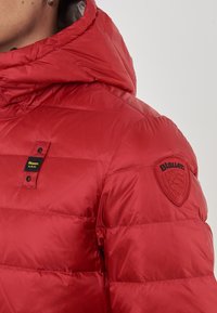 Primo piano di una giacca trapuntata rossa con cappuccio, etichetta nera e gialla "Blauer U.S.A." sul petto e toppa rossa "Blauer" sulla manica superiore.
