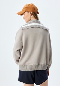 Jasnoszara sweter z prążkowanym kołnierzem i dołem, z paskowanym akcentem na kołnierzu. Połączony z ciemnymi szortami i brązową czapką.