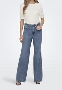 Höga, ljusblå denimjeans med vida ben, med bälte i midjan och subtila blekningar, tillsammans med en texturerad krämfärgad topp.