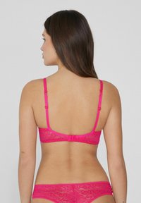 Růžový krajkový bralette s nastavitelnými ramínky, zapínáním na háček a očko na zadní straně a květinovým vzorem. Přiléhá těsně k tělu.