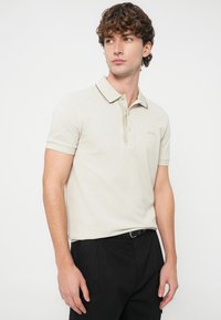 Mladý muž v svetlobéžovom polotričku s čiernym lemom na golieri a čiernych nohaviciach stojaci pred jednoduchým bielym pozadím.