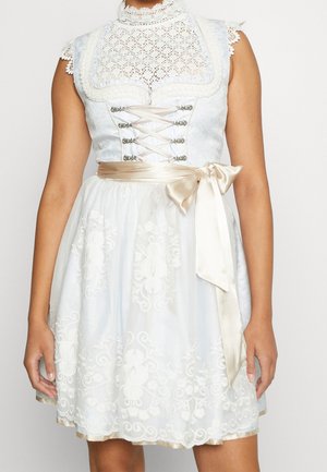 Modèle portant une robe dirndl blanche en dentelle, avec un corsage de style corset, une jupe brodée et une écharpe en satin couleur champagne nouée à la taille.