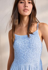 Robe bleu clair avec corsage froncé et bretelles spaghetti. Le tissu a une texture douce et se déploie en une jupe légèrement plissée.