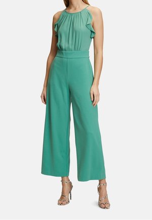 Kvinde iført en ærmeløs teal jumpsuit med flæsebind, bukser med vidde og højhælede sandaler i slangeskind på hvid baggrund.