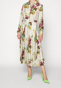 Žena ve volných rukávech, skládací květinové šaty s fialovými a modrými květy, doplněné jasně zelenými botami na vysokém podpatku s špičatou špičkou.